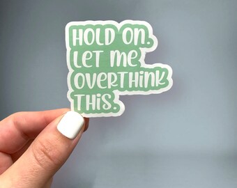 May include: A sticker with the text "HOLD ON. LET ME OVERTHINK THIS." in a green, bubble-style font. The sticker has a white border and is held by a hand with a white nail polish.