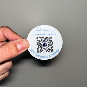 May include: A white circular sticker with the text "Adriana's Bachelorette Party" in a script font. A QR code is in the center, with the words "BUY THE BRIDE A DRINK!" around the bottom edge. The sticker is held by a hand against a gray background.