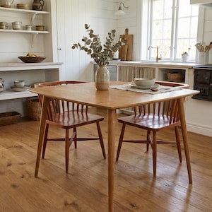 Op de afbeelding: Een vierkante, lichtbruine houten eettafel met afgeronde hoeken en vier taps toelopende poten, vergezeld door twee bijpassende houten stoelen met spijlenrug. In het midden van de tafel staat een keramische vaas met groene eucalyptus.
