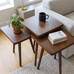 Peut inclure: Un ensemble de trois tables gigognes en bois brun foncé avec des coins arrondis et des pieds fuselés. Une plante Pilea verte dans un pot en terre cuite est posée sur la plus petite table, tandis qu'une tasse noire et trois livres sont posés.