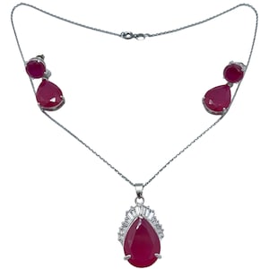 Puede incluir: Un collar y pendientes de plata con un colgante en forma de lágrima y pendientes a juego. El colgante y los pendientes presentan una gran piedra roja rodeada de piedras transparentes más pequeñas.