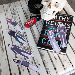 May include: A set of paper bookmarks featuring characters from the TV show Bones. The bookmarks are printed with the characters' names and the show's title. The bookmarks are on a wooden surface with a book and a belt buckle.