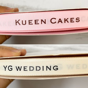 May include: Two rolls of ribbon with different colors and text. The top ribbon is pink with the text "KUEEN CAKES" printed in brown. The bottom ribbon is cream with the text "YG WEDDING" printed in black.