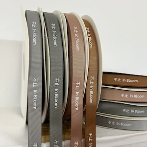 May include: A set of eight spools of ribbon in various shades of brown and gray. Each spool has the text "In Bloom" printed in white on the ribbon.