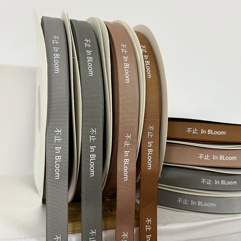 May include: A set of eight spools of ribbon in various shades of brown and gray. Each spool has the text "In Bloom" printed in white on the ribbon.