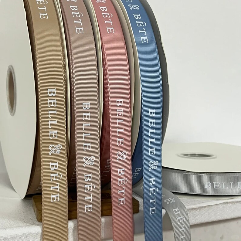 May include: Five spools of ribbon in different colors: beige, brown, pink, blue, and gray. Each spool has the text "BELLE & B&Ecirc;TE" printed on it.
