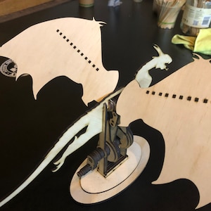 Könnte beinhalten: Ein hölzernes Drachenmodell mit einem detaillierten, komplizierten Design. Der Drache befindet sich in einer Flugpose mit ausgebreiteten Flügeln. Das Modell ist aus hellbraunem Holz gefertigt und hat eine dunkelbraune Oberfläche.