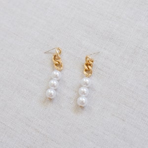 May include: A pair of gold-toned chain earrings with three white faux pearls dangling from each chain. The earrings are displayed on a neutral, light-colored surface.