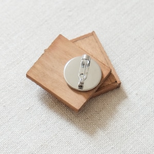 May include: A wooden pin with a round metal backing and a safety pin closure. The pin is made of two pieces of wood that are joined together.