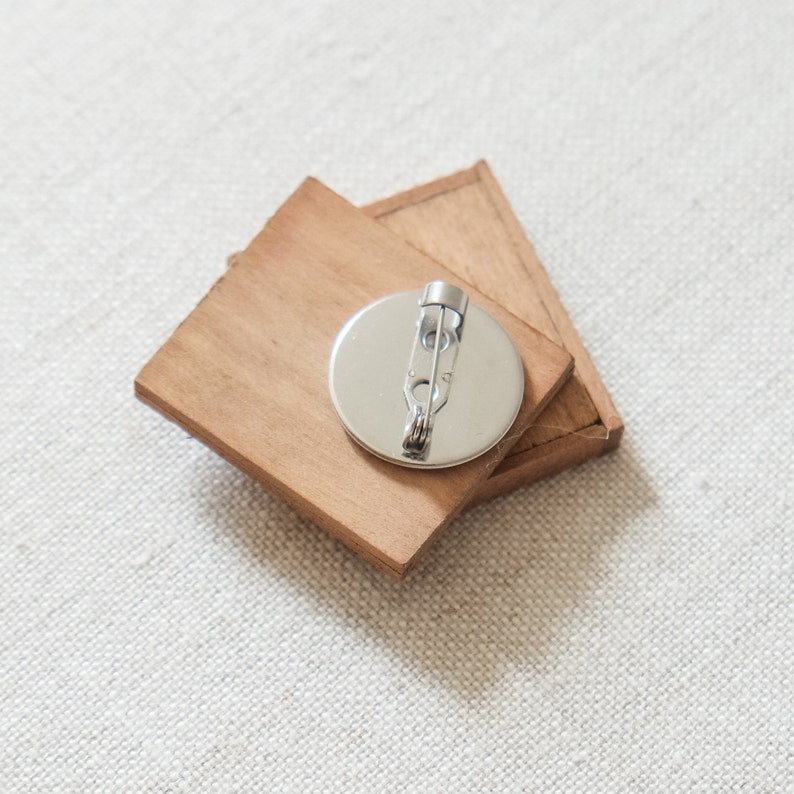 May include: A wooden pin with a round metal backing and a safety pin closure. The pin is made of two pieces of wood that are joined together.