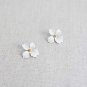 Puede incluir: Un par de pendientes de flores blancas con centros dorados. Los pendientes están sobre un fondo de tela blanca.