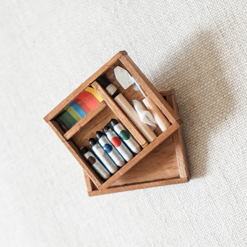 May include: A wooden box with a hinged lid, containing a set of colorful paint tubes, a paint scraper, and a paintbrush. The box is open, revealing the contents.