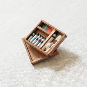 May include: A wooden box with a hinged lid, containing a set of colored pencils, a white eraser, and a wooden pencil. The box is open and the contents are visible.