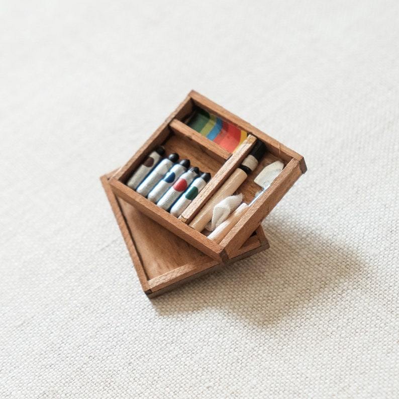 May include: A wooden box with a hinged lid, containing a set of colored pencils, a white eraser, and a wooden pencil. The box is open and the contents are visible.
