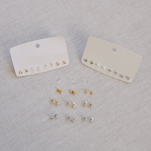 May include: Two white cards with a variety of silver and gold stud earrings displayed on them. The earrings are arranged in rows and include different shapes, such as circles, triangles, and hearts. The earrings are all small and delicate.