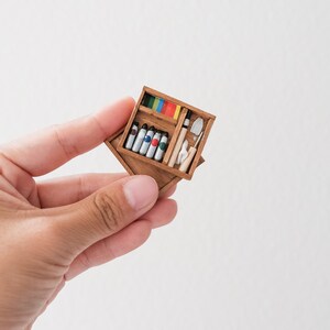 May include: A wooden box with a hinged lid, containing a set of paint tubes, a small trowel, and a paintbrush. The box is open and the contents are visible.
