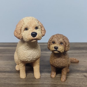 Op de afbeelding: Twee keramische hondenbeeldjes, een lichtbruin en een donkerbruin, zitten op een houten oppervlak. Beide honden hebben zwarte ogen en neuzen.