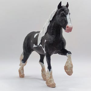 Pode incluir: Figurinha de cavalo de brinquedo preto e branco com crina e cauda brancas. O cavalo tem cascos marrons e está em pé sobre uma superfície branca.