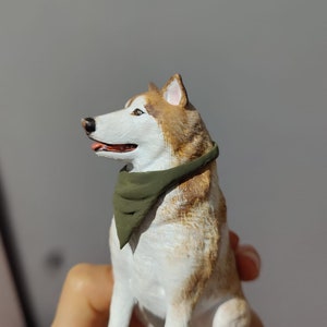 May include: A white and brown figurine of a husky dog wearing a green bandana. The dog is sitting with its head turned to the side and its mouth slightly open.
