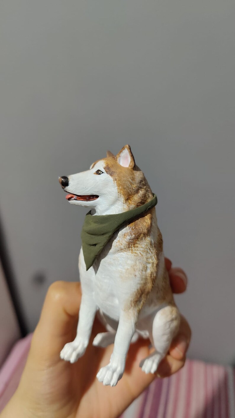 May include: A white and brown figurine of a husky dog wearing a green bandana. The dog is sitting with its head turned to the side and its mouth slightly open.