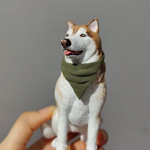 May include: A white and brown husky dog figurine wearing a green bandana. The figurine is sitting up and looking to the right. The figurine is being held in a person's hand.