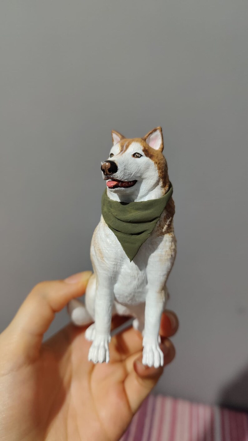 May include: A white and brown husky dog figurine wearing a green bandana. The figurine is sitting up and looking to the right. The figurine is being held in a person's hand.