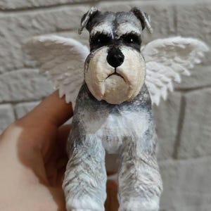 Op de afbeelding: Handgeschilderd beeldje van een Schnauzer-hond met witte engelen vleugels. De hond heeft een grijs-witte vacht, een zwarte neus en een serieuze uitdrukking. De vleugels zijn wit en gedetailleerd, wat een grillige toets toevoegt.