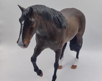 May include: A brown and black horse figurine with white markings on its face and legs. The horse is standing with its head turned to the side and its legs slightly bent.
