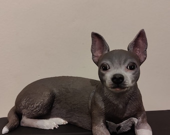 May include: A gray and white figurine of a small dog lying down. The dog has large ears and a short snout.
