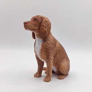 May include: A brown and white dog figurine sitting with its head tilted up. The figurine is made of a textured material that resembles fur.