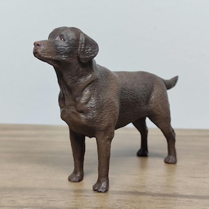 May include: A brown Labrador Retriever dog figurine. The figurine is standing on a wooden surface and is looking to the left.