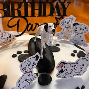 Op de afbeelding: Een witte taart versierd met zwarte pootafdrukken en Dalmatische themaversieringen. De taart heeft een zwarte "BIRTHDAY Darcy" taarttopper en verschillende Dalmatische hondenuitsparingen. Een 3D Dalmatische hondenfiguur staat erop.