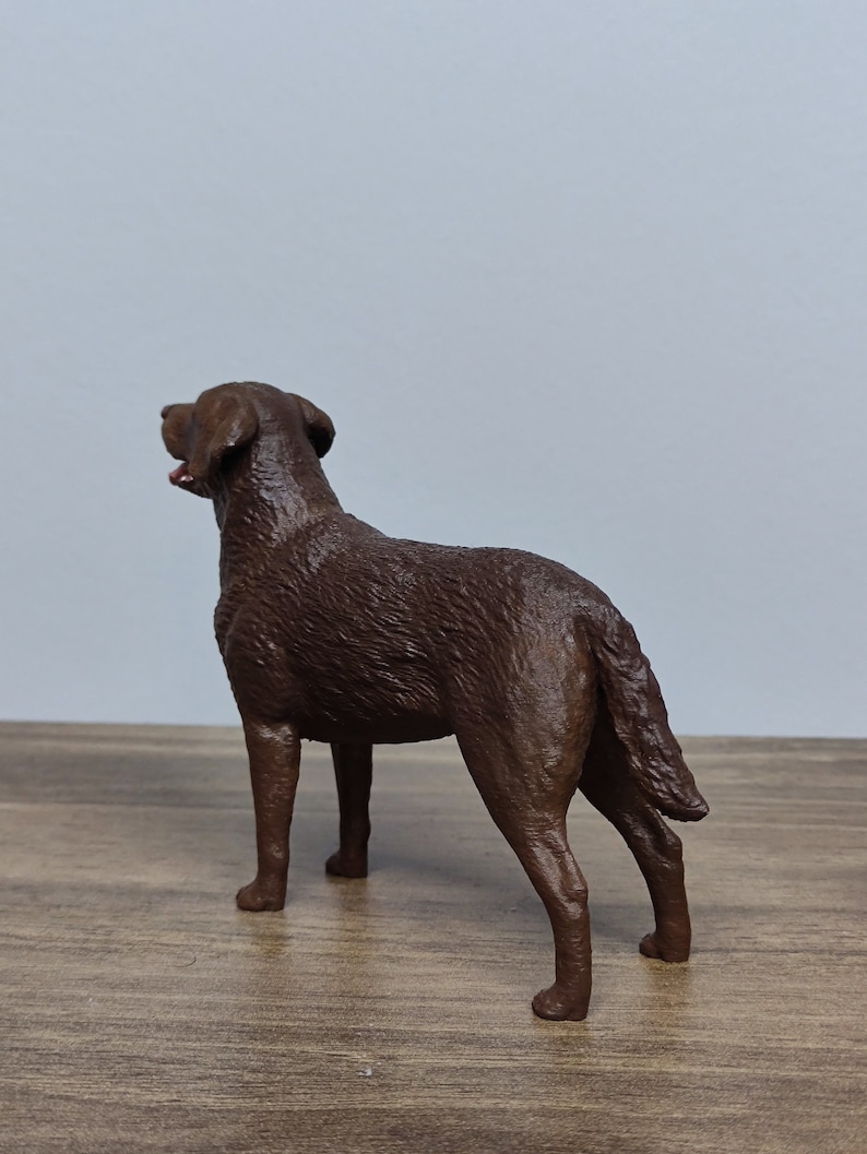 May include: A chocolate brown Labrador Retriever dog figurine. The figurine is standing on a wooden surface and is looking up.