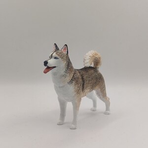May include: A hand-painted figurine of a Siberian Husky dog. The dog is standing on a white surface and is looking to the left. The figurine is painted in shades of white, gray, and brown.