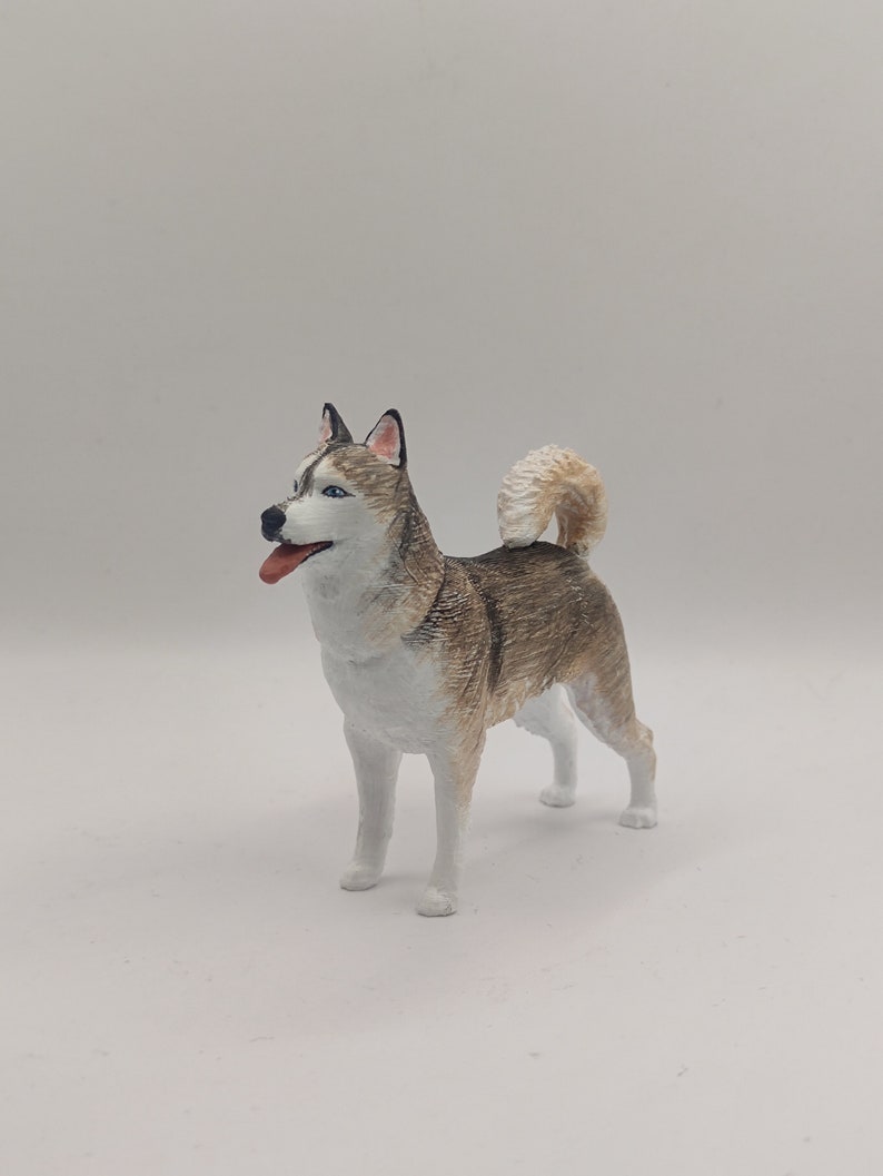 May include: A hand-painted figurine of a Siberian Husky dog. The dog is standing on a white surface and is looking to the left. The figurine is painted in shades of white, gray, and brown.