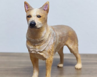 May include: A brown and white Australian Cattle Dog figurine with pink ears. The figurine is standing on a wooden surface.