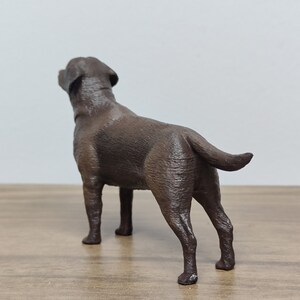 May include: A brown Labrador Retriever dog figurine standing on a wooden surface. The dog is looking up and has its tail curled.