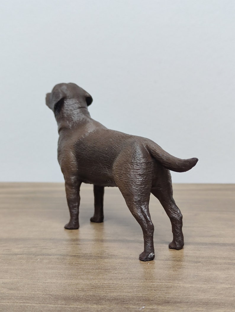 May include: A brown Labrador Retriever dog figurine standing on a wooden surface. The dog is looking up and has its tail curled.