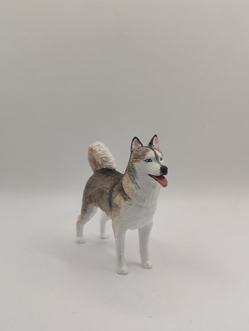May include: A white, gray, and brown figurine of a husky dog with blue eyes. The dog is standing on a white surface and has its tongue sticking out.