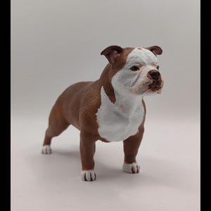 May include: A brown and white bulldog figurine with a white patch on its chest and white markings on its face. The figurine is standing on a white surface.