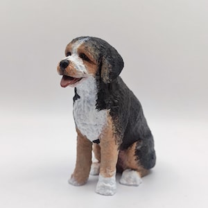Pode incluir: Uma pequena figura de cão preto, marrom e branco sentada com a cabeça virada para o lado. A figura é feita de cerâmica pintada.
