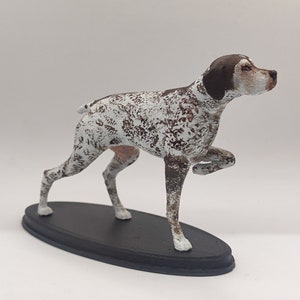 May include: A brown and white figurine of a German Shorthaired Pointer dog standing on a black oval base. The dog is posed in a running position with its right leg raised.