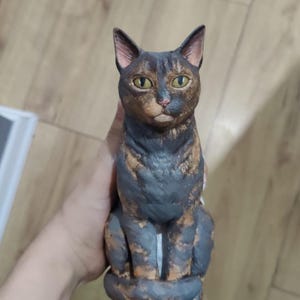 May include: A ceramic cat figurine with brown, black, and orange markings. The cat is sitting with its paws tucked underneath it and has a serious expression on its face.