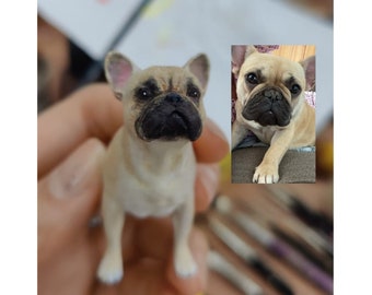 May include: A small, beige French bulldog figurine with black eyes and nose. The figurine is standing on a hand, with a blurred background.