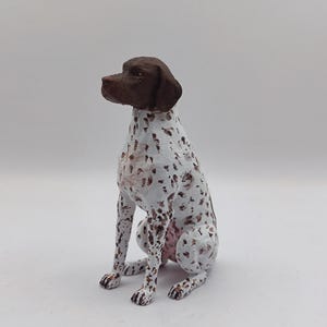 May include: A brown and white German Shorthaired Pointer figurine sitting on a white surface. The figurine is painted with realistic details, including brown fur, white spots, and a pink nose.