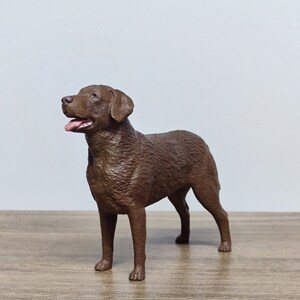May include: A chocolate brown Labrador Retriever dog figurine. The figurine is standing on a wooden surface and is looking up and to the left.