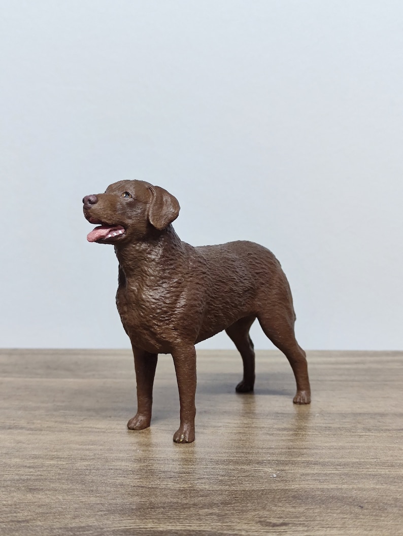 May include: A chocolate brown Labrador Retriever dog figurine. The figurine is standing on a wooden surface and is looking up and to the left.