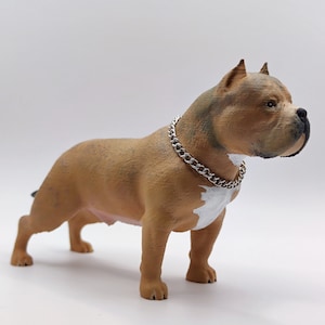 Könnte beinhalten: Eine braune und weiße American Bully Hundefigur mit einem silbernen Kettenhalsband. Die Figur steht auf einer weißen Oberfläche.
