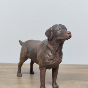 May include: A brown Labrador Retriever dog figurine. The figurine is standing on a wooden surface and is looking up.