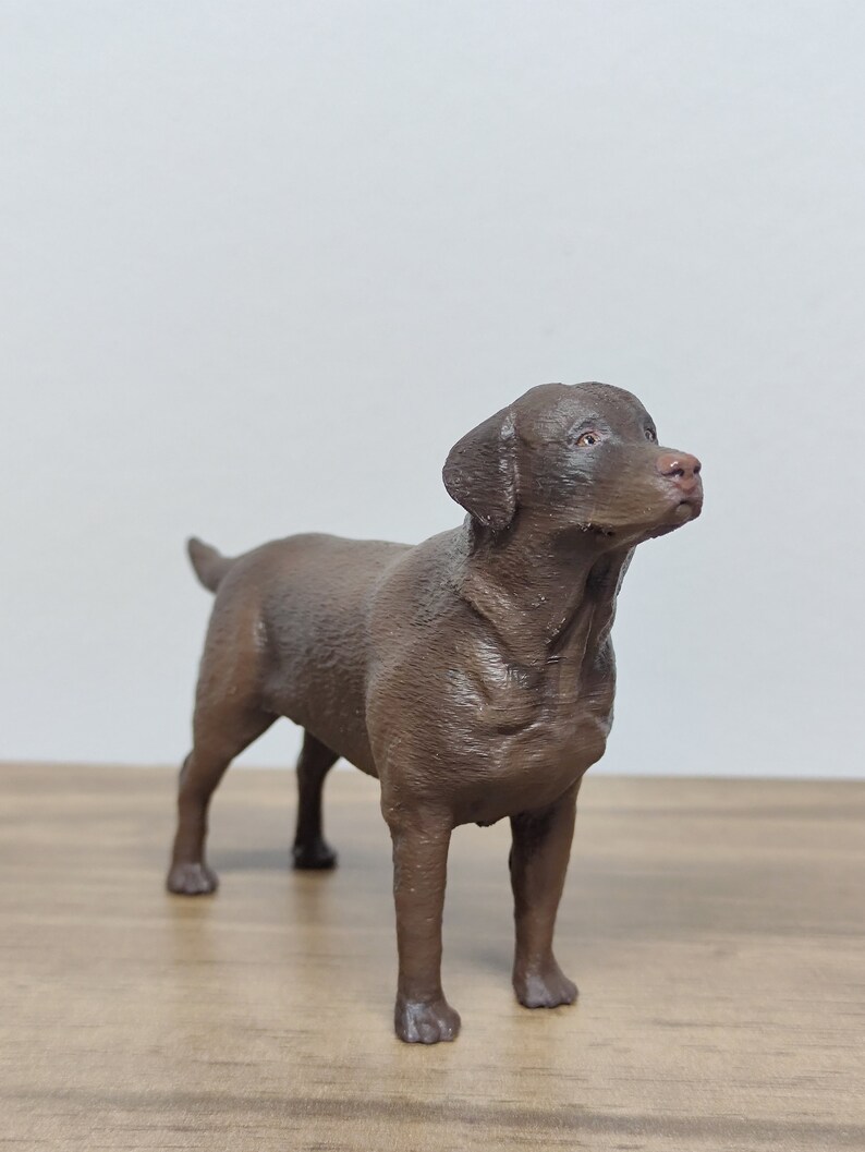 May include: A brown Labrador Retriever dog figurine. The figurine is standing on a wooden surface and is looking up.